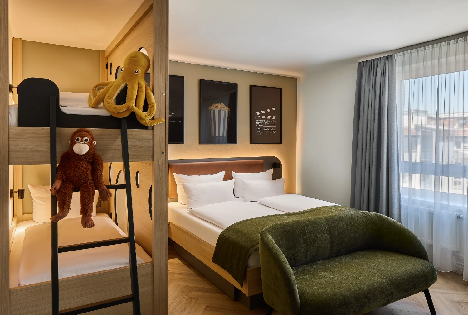 Modernes Familienzimmer mit Doppelbett und integriertem Etagenbett. Auf dem Etagenbett sitzen ein Plüschaffe und ein Kuschelkrake. Heller Wohnbereich mit Sessel, großem Fenster und gemütlicher Atmosphäre.