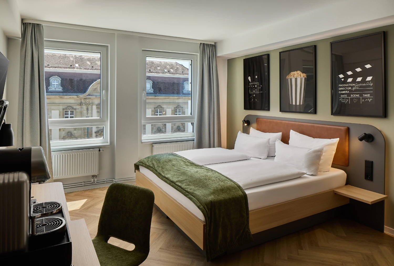 Modernes Hotelzimmer mit komfortablem Doppelbett, Schreibtisch und Kaffeemaschine. Große Fenster sorgen für viel Tageslicht und bieten einen Blick auf die historische Karlsruher Innenstadt.