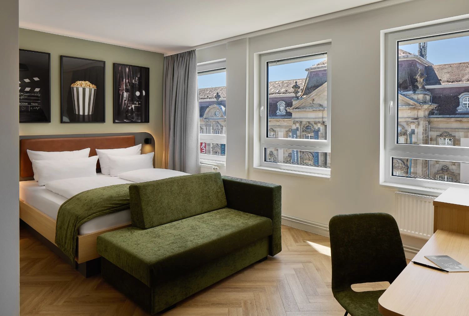 Modernes Hotelzimmer mit komfortablem Doppelbett, gemütlichem Sofa und Schreibtisch. Große Fenster sorgen für viel Tageslicht und bieten einen Blick auf die historische Karlsruher Innenstadt.