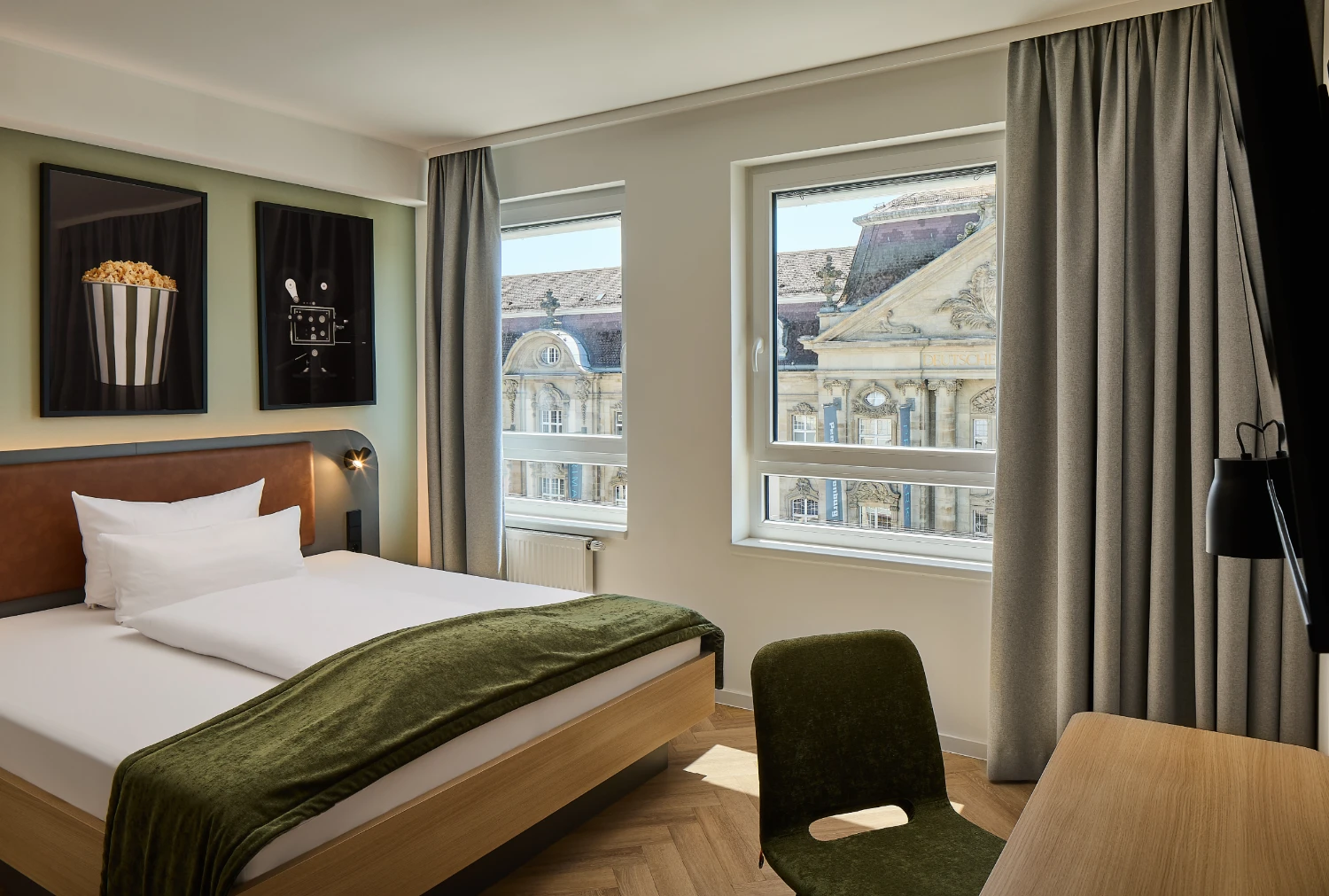 Modernes Hotelzimmer mit komfortablem Doppelbett, gemütlichem Sofa und Schreibtisch. Große Fenster sorgen für viel Tageslicht und bieten einen Blick auf die historische Karlsruher Innenstadt.