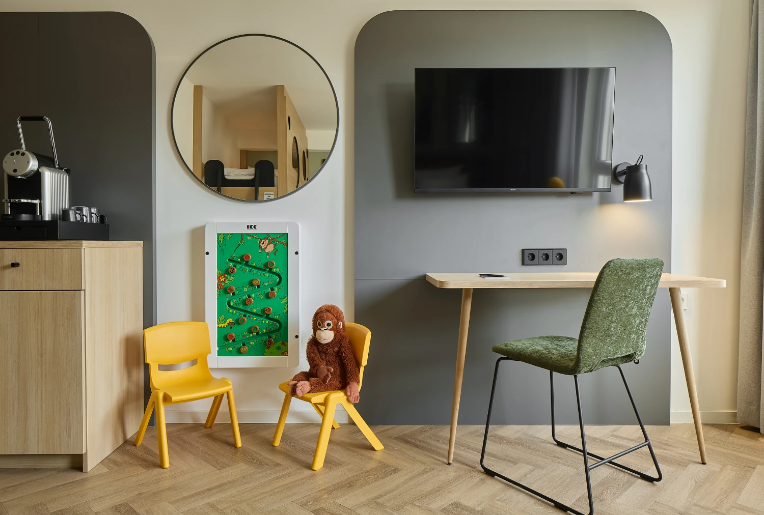 Familienfreundlicher Bereich eines modernen Hotelzimmers mit Schreibtisch, Smart-TV, Kaffeemaschine sowie einer Spielecke mit zwei Kinderstühlen, Plüschaffe und Wandspiel. Im Spiegel ist ein Etagenbett zu sehen.