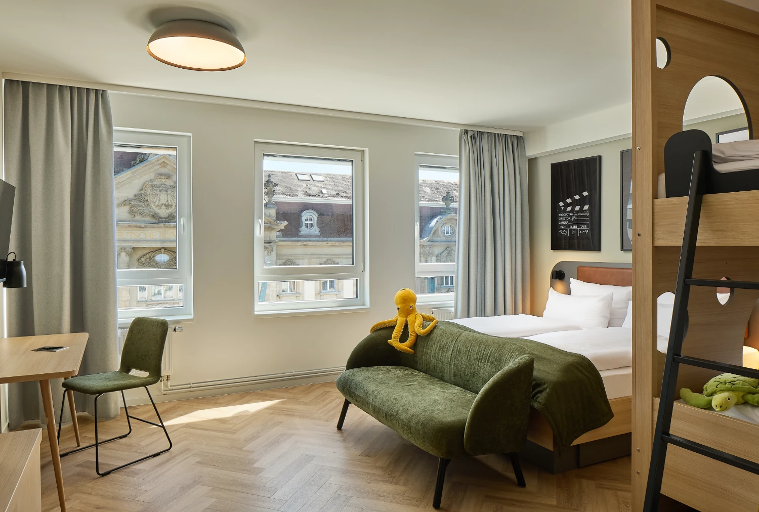 Helles Familienzimmer mit Doppelbett, integriertem Etagenbett, gemütlichem Sofa und Schreibtisch. Große Fenster sorgen für viel Tageslicht und bieten Blick auf die historische Karlsruher Innenstadt.