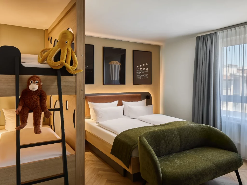 Modernes Familienzimmer mit Doppelbett und integriertem Etagenbett. Auf dem Etagenbett sitzen ein Plüschaffe und ein Kuschelkrake. Heller Wohnbereich mit Sessel, großem Fenster und gemütlicher Atmosphäre.