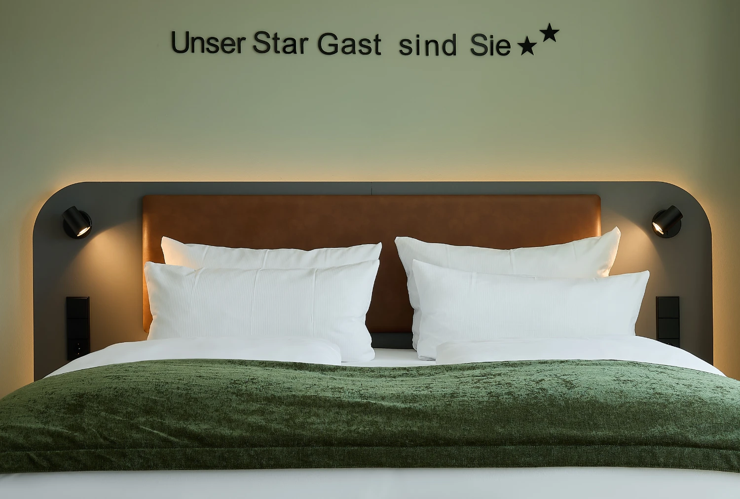 Nahaufnahme eines komfortablen Doppelbetts mit indirekter Beleuchtung, Leselampen und hochwertiger Bettwäsche. Über dem Kopfteil steht der Schriftzug „Unser Star Gast sind Sie“ und unterstreicht die persönliche Wohlfühlatmosphäre.