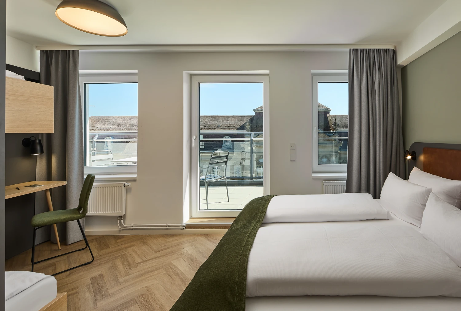 Helles Familienzimmer mit Doppelbett und Schreibtisch. Große Fenster sorgen für viel Tageslicht und bieten Blick auf den Balkon und die historische Karlsruher Innenstadt.