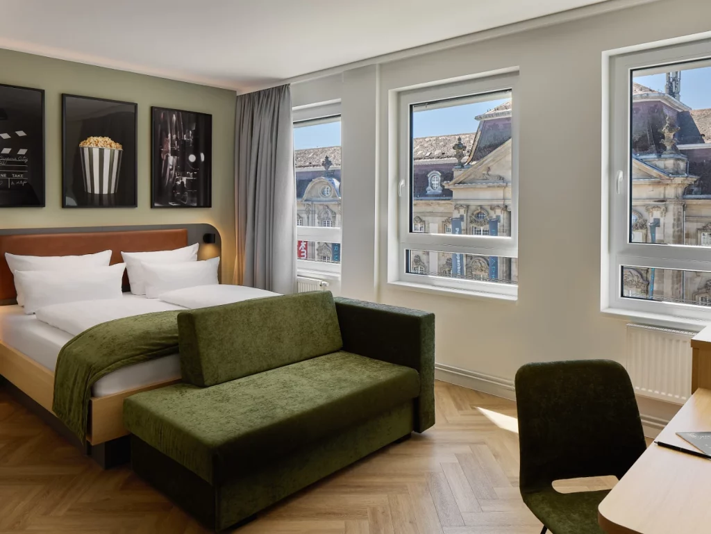Modernes Hotelzimmer mit komfortablem Doppelbett, gemütlichem Sofa und Schreibtisch. Große Fenster sorgen für viel Tageslicht und bieten einen Blick auf die historische Karlsruher Innenstadt.