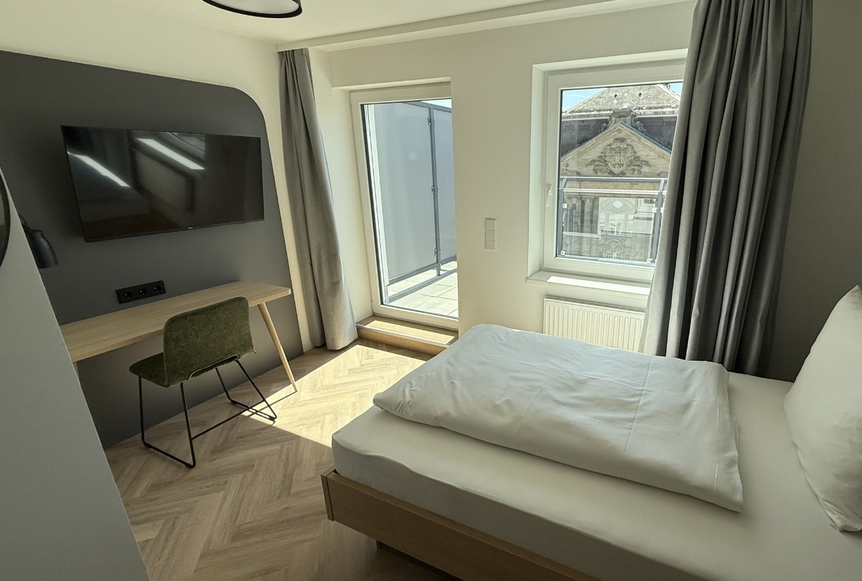 Helles Einzelzimmer mit großem Fenster und Blick auf die Stadt, ausgestattet mit Bett, Schreibtisch und Stuhl