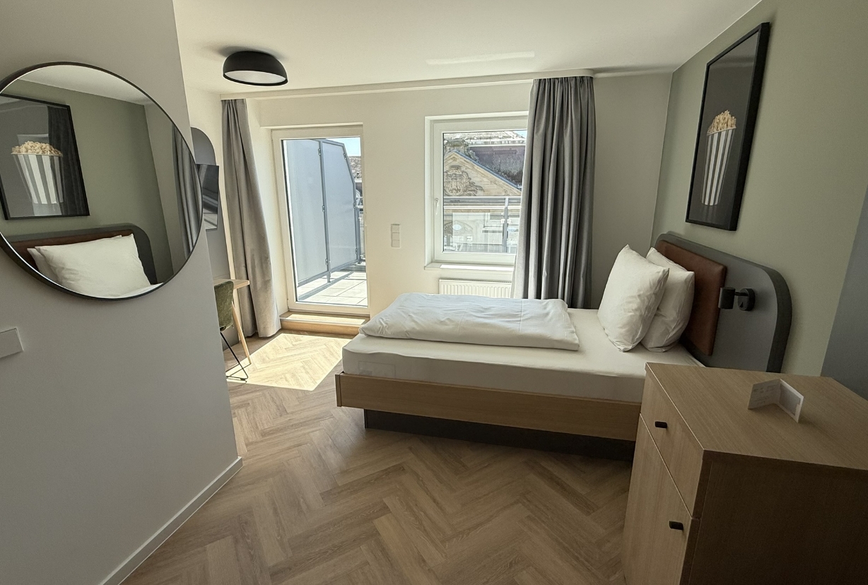 Helles Einzelzimmer mit Holzfußboden, Schreibtisch und Zugang zum Balkon