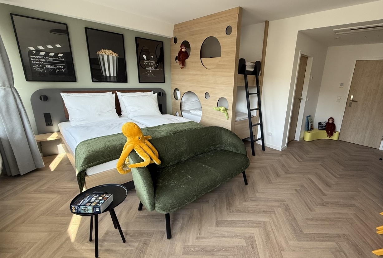 Familienzimmer mit Doppelbett, Kinderstockbett mit Kletterleiter, grünem Sofa, Spieltisch und verspielter Dekoration im Cinema City Hotel