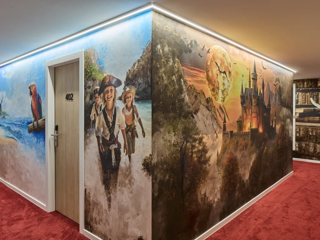 Thematisch gestalteter Hotelflur mit großflächigem Wandmotiv, rotem Teppich und beleuchteten Zimmerzugängen. Das moderne Design greift das Kino- und Filmkonzept des Hotels eindrucksvoll auf.