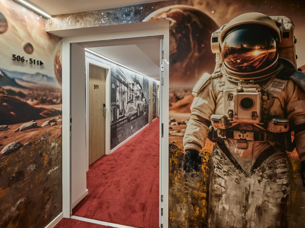 Thematisch gestalteter Hotelflur mit großflächigem Wandmotiv, rotem Teppich und beleuchteten Zimmerzugängen. Das moderne Design greift das Kino- und Filmkonzept des Hotels eindrucksvoll auf.