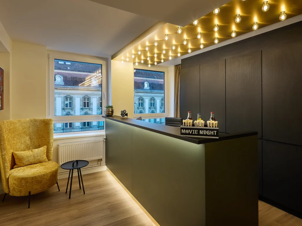 Moderne Hotelrezeption mit stilvollem Empfangstresen, gemütlichem Loungesessel und dekorativer Beleuchtung im Kino-Stil. Große Fenster sorgen für Tageslicht und bieten einen Blick auf die Karlsruher Innenstadt.